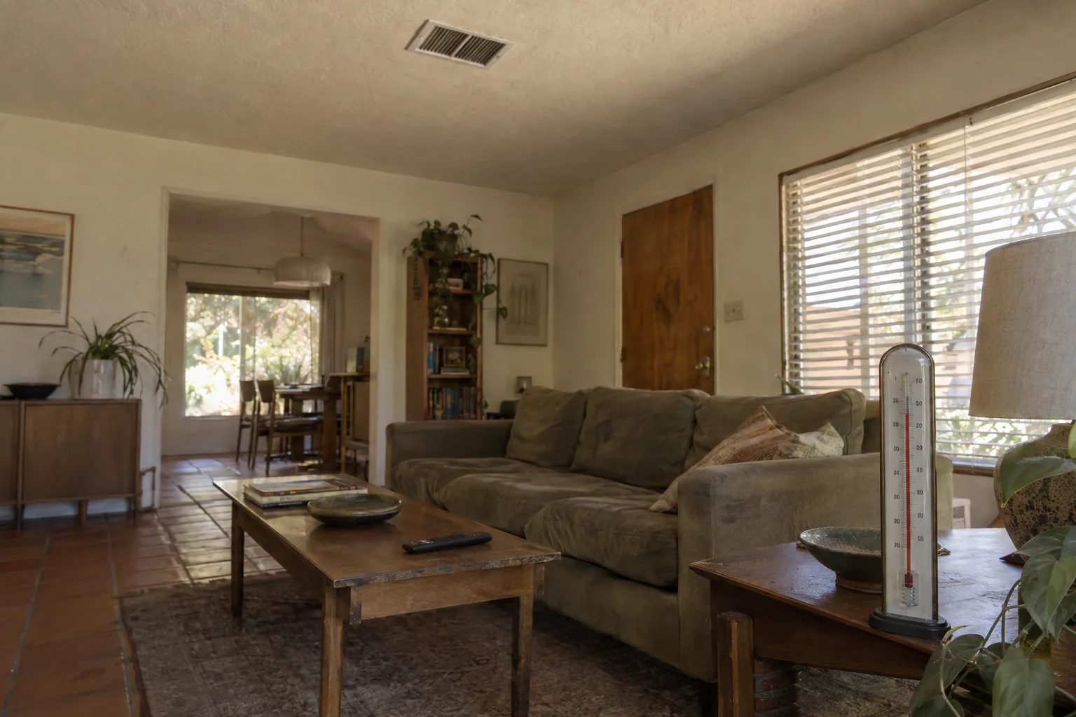 Sun-drenched living room interior of a mid-century ranch house with stucco walls and terracotta tile floor, warm light filtering through blinds, a ceiling vent visible above, and a thermometer on a side table suggesting the room is uncomfortably hot despite the air conditioning running