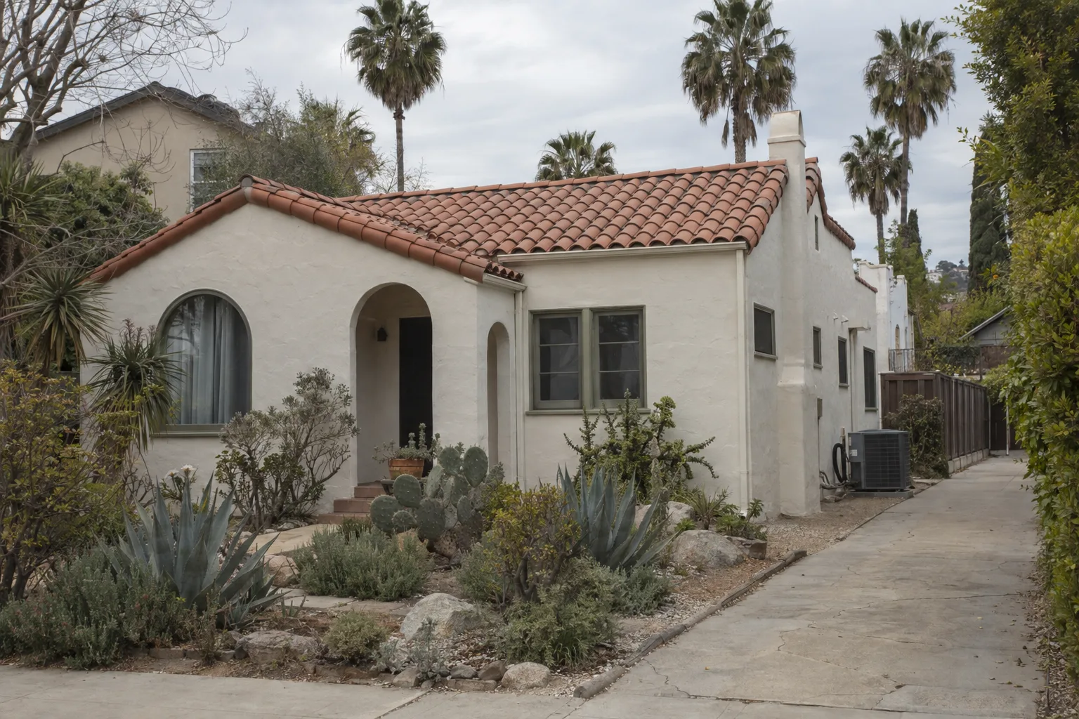 A single-story Spanish-style stucco house in Los Angeles with a tile roof, drought-tolerant front yard landscaping, palm trees in the background, and an AC condenser unit visible along the side of the house