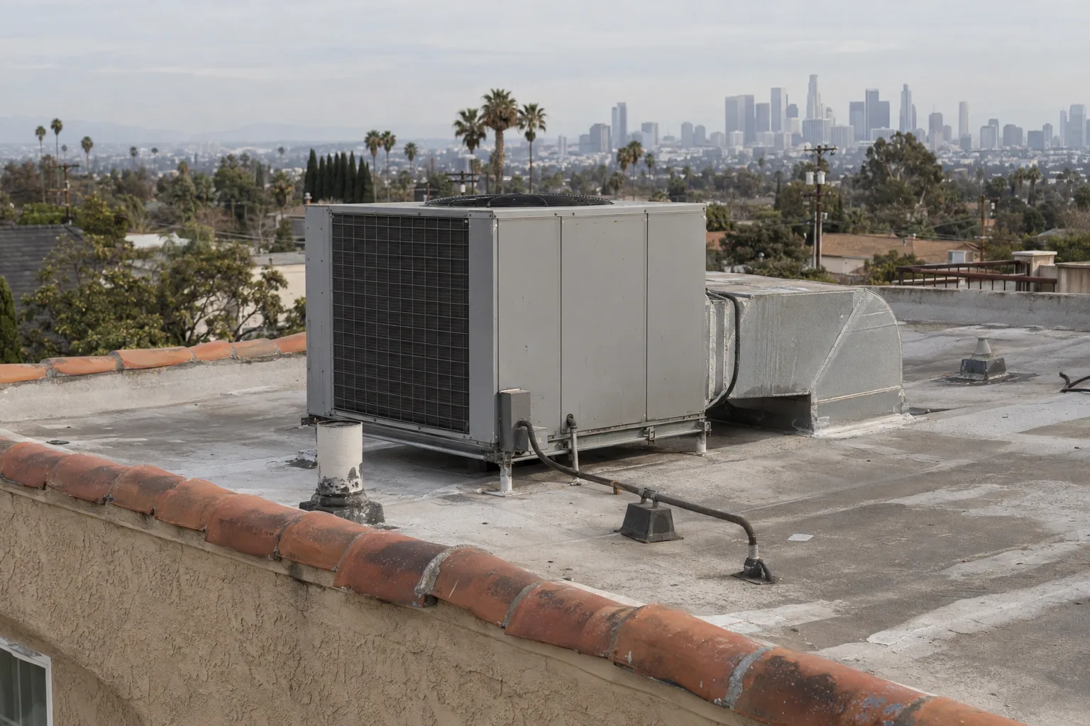A new packaged AC rooftop unit sitting on a flat stucco residential roof with a terracotta parapet, clean ductwork penetrations, and a hazy Los Angeles skyline faintly visible in the background under warm late-afternoon light.