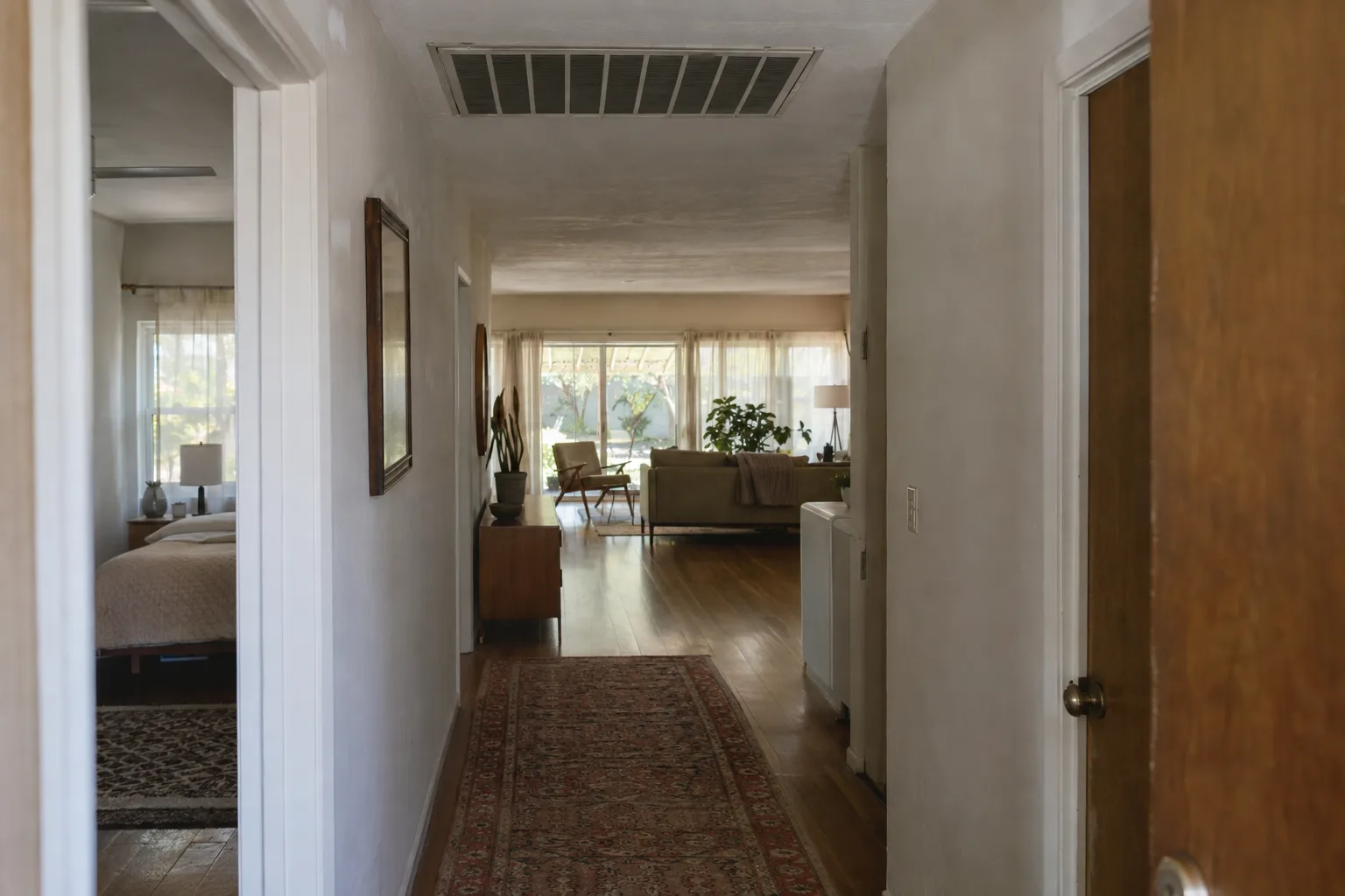 A sunlit hallway in a mid-century ranch house looking toward an open living room, with a ceiling return-air grille visible above, illustrating how a central HVAC system connects every room in the home