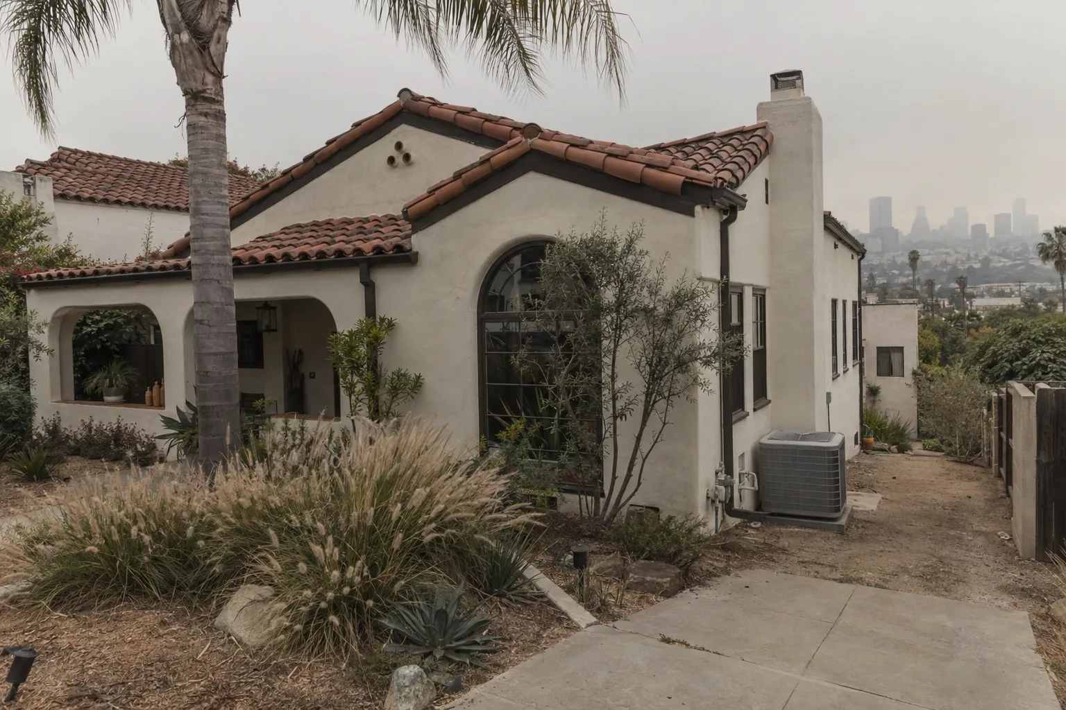 A Spanish-style stucco house in Los Angeles photographed in late afternoon light, with a central air condenser unit along the side yard, a palm tree in the foreground, and a subtly hazy sky