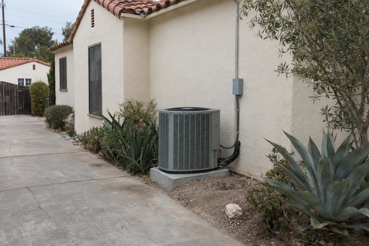 Side yard of a Spanish-style stucco Los Angeles home with a central air conditioning condenser unit sitting on a concrete pad beside the exterior wall, terracotta tile roof and drought-tolerant plants visible in the background