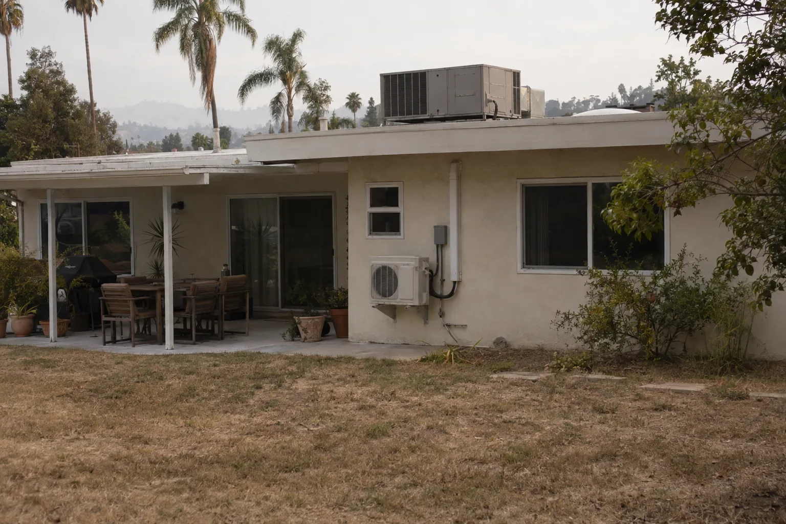 Rear exterior of a flat-roofed mid-century ranch stucco home in Los Angeles with a rooftop packaged AC unit visible above the parapet and a small mini-split outdoor compressor mounted on the back wall, palm trees and dry landscaping in the background