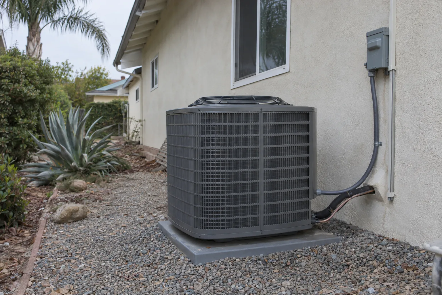 A freshly installed central air conditioning condenser unit on a gravel pad beside a cream stucco ranch house, with copper refrigerant lines strapped neatly to the exterior wall and drought-tolerant plants in the foreground under bright Los Angeles afternoon light.