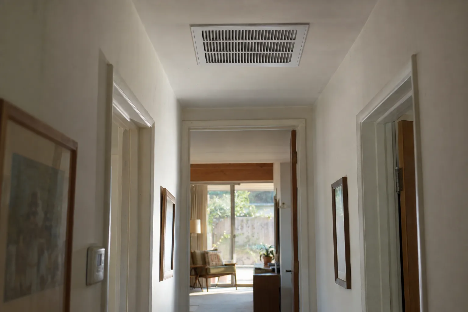 A white ceiling supply vent flush-mounted in the smooth drywall ceiling of a mid-century ranch house hallway, with warm California afternoon light coming through a window at the end of the corridor.
