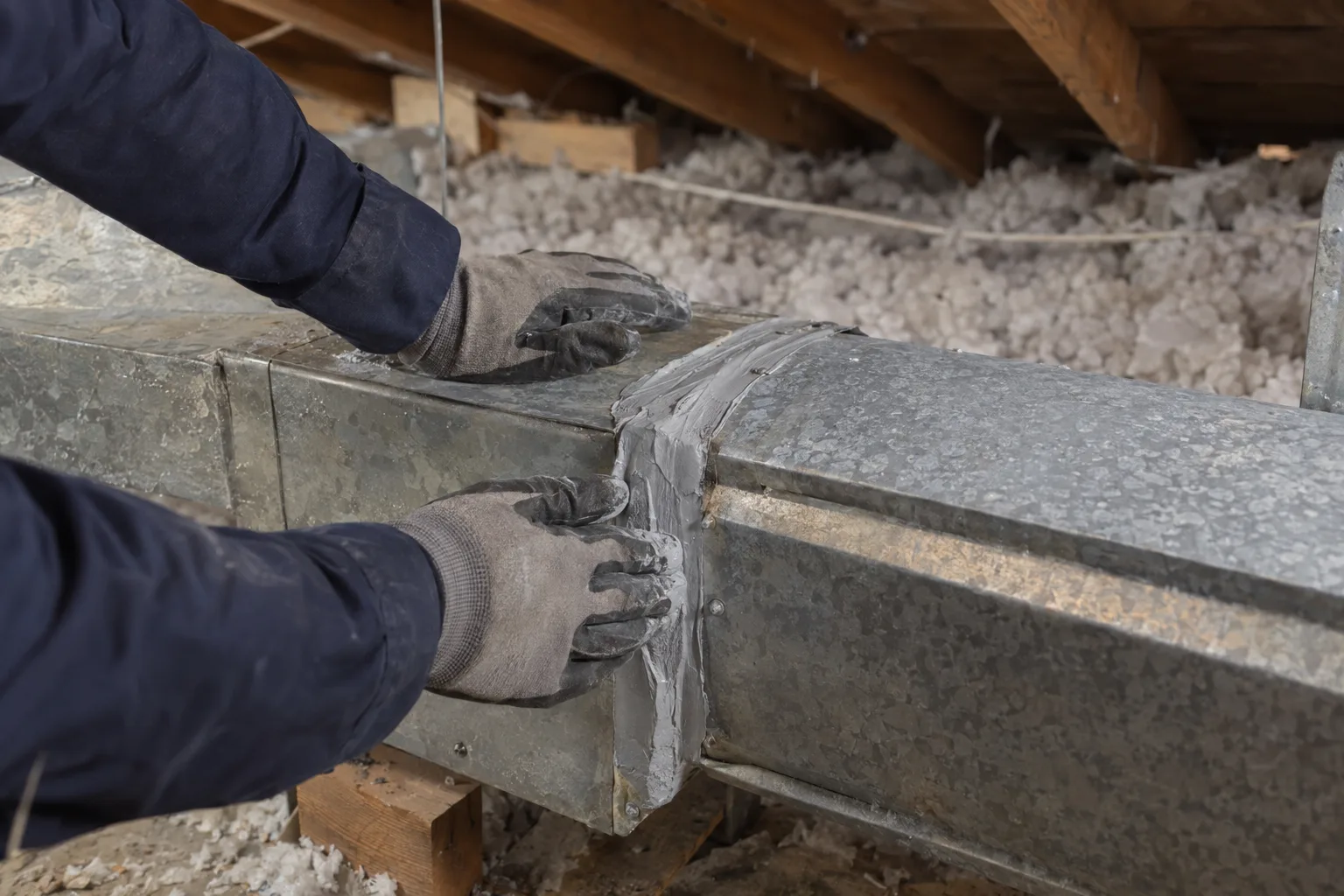 Gloved hands applying mastic sealant to a leaking sheet-metal duct joint inside a sunlit Los Angeles attic with timber rafters and loose insulation visible in the background