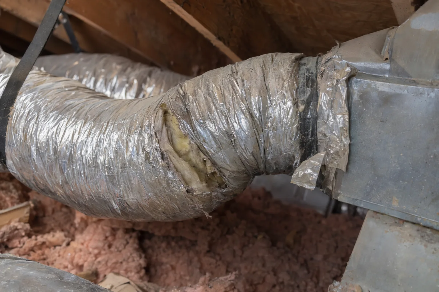 A section of silver flexible duct in a residential attic showing a torn outer jacket, a sharp kink, and peeling foil tape at a duct boot seam, lit by warm natural light from a nearby attic vent