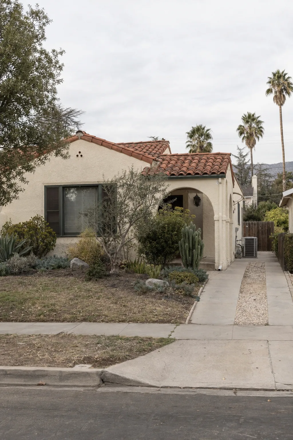A single-story stucco Spanish-style home in a San Gabriel Valley neighborhood at late afternoon, with terracotta tile roof, drought-tolerant front yard, and a side-yard HVAC condenser visible along the fence.