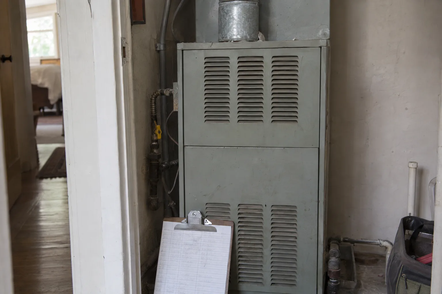 An aging upright gas furnace installed in a residential utility space, with a clipboard of inspection notes leaning against it in warm indoor light