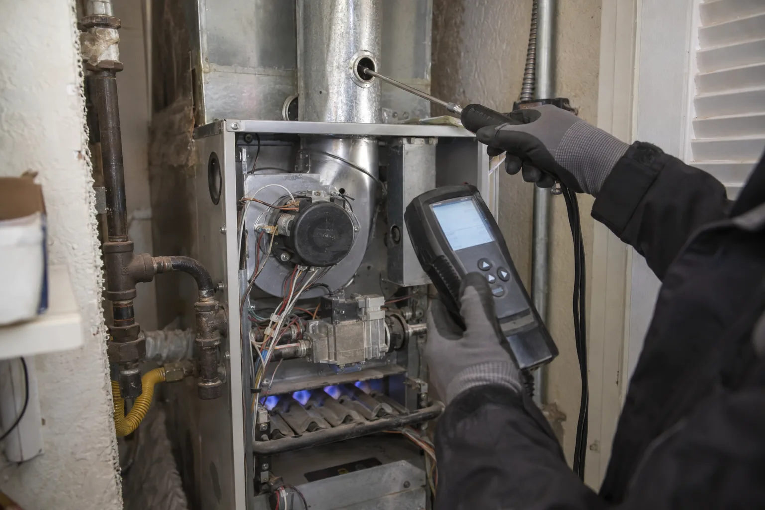 A gas furnace cabinet open in a residential utility closet, with a technician's gloved hands positioning a combustion analyzer probe into the flue port during a tune-up inspection.