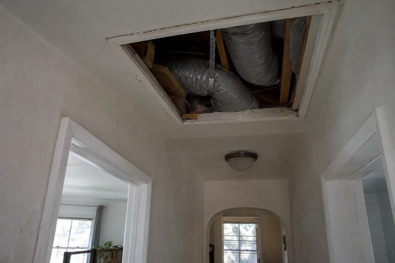 Open ceiling access panel in a Los Angeles stucco home revealing flexible grey duct runs between wooden joists, photographed from a low hallway angle in afternoon light