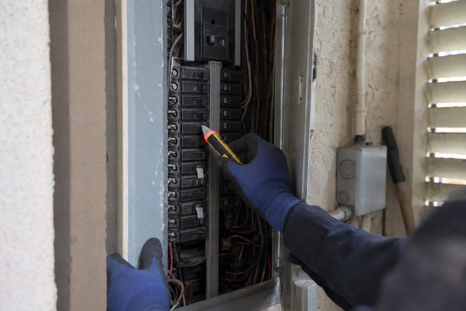 A technician's gloved hands holding a voltage tester near an open residential electrical panel inside a utility closet