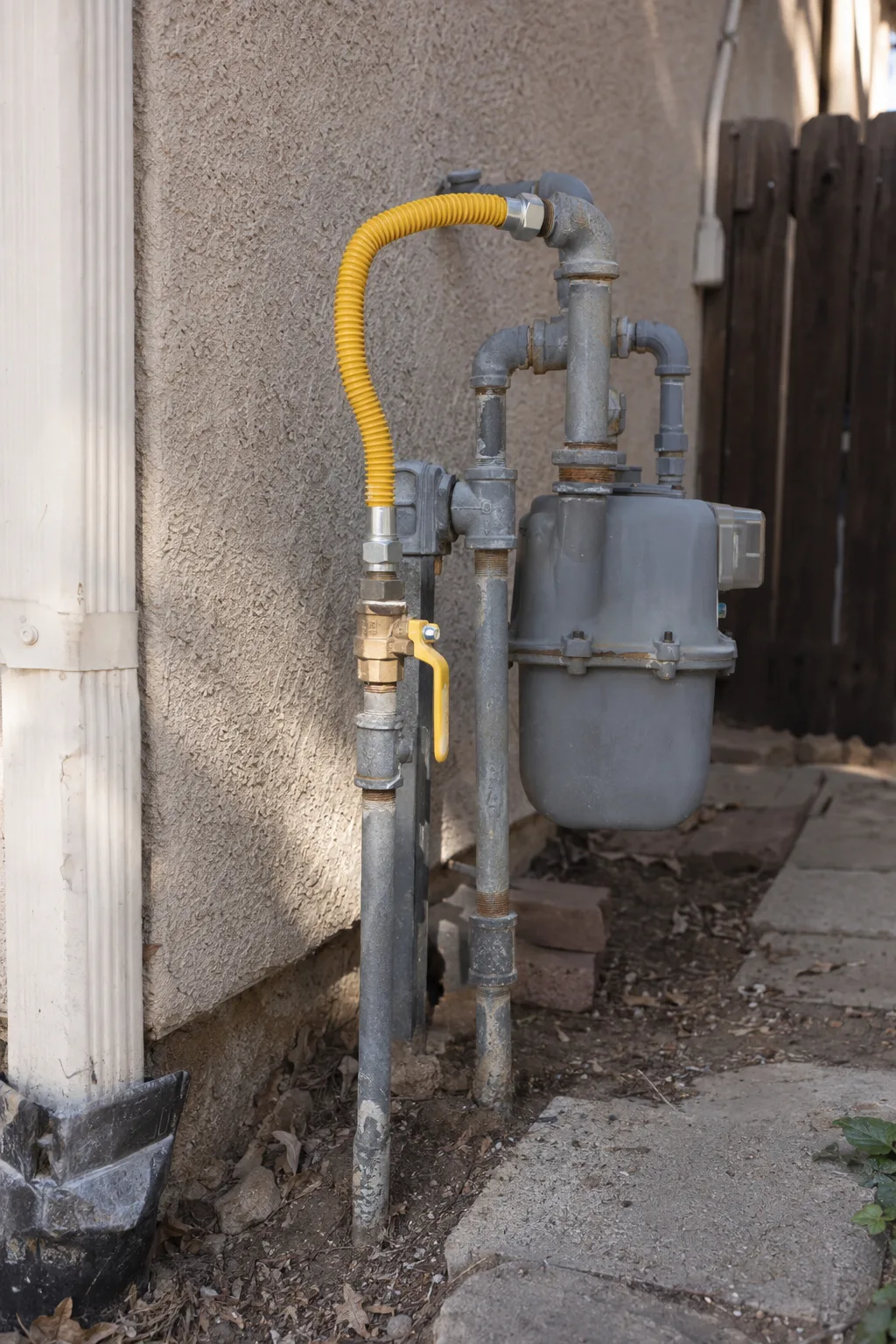 A yellow flexible gas supply line and shut-off valve mounted on a stucco exterior wall in a residential side yard