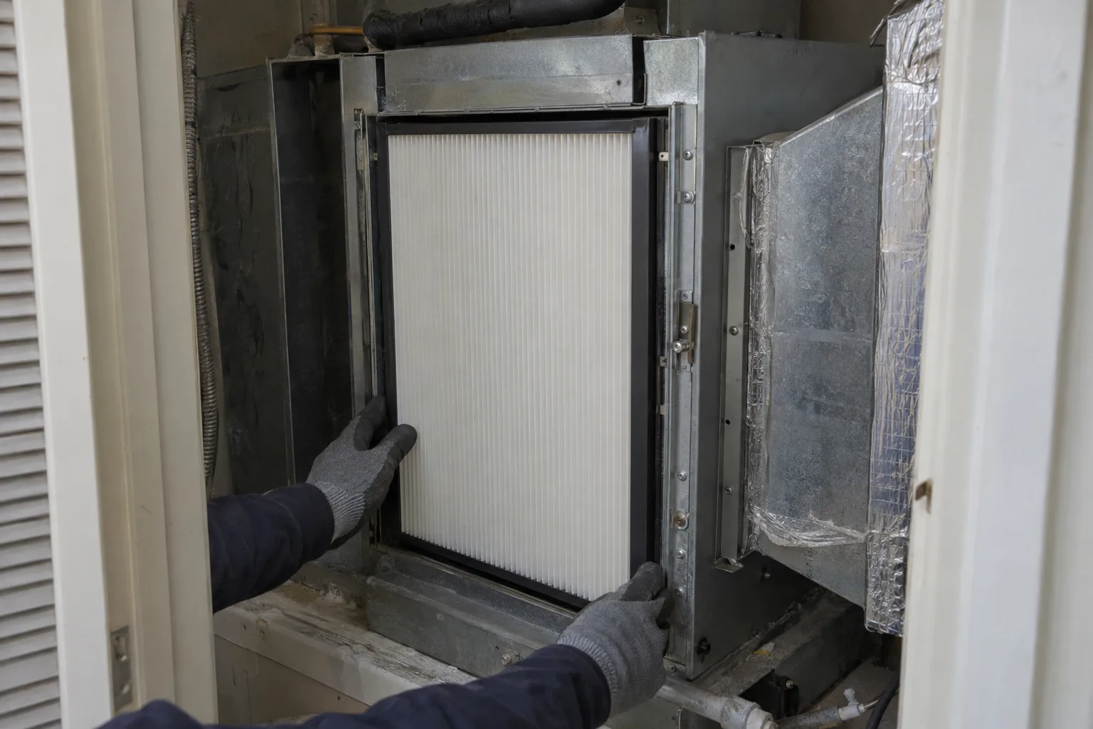 Pleated HEPA filter cartridge correctly seated inside a residential air handler housing in a utility closet, with gloved hands visible at the edges staging the unit