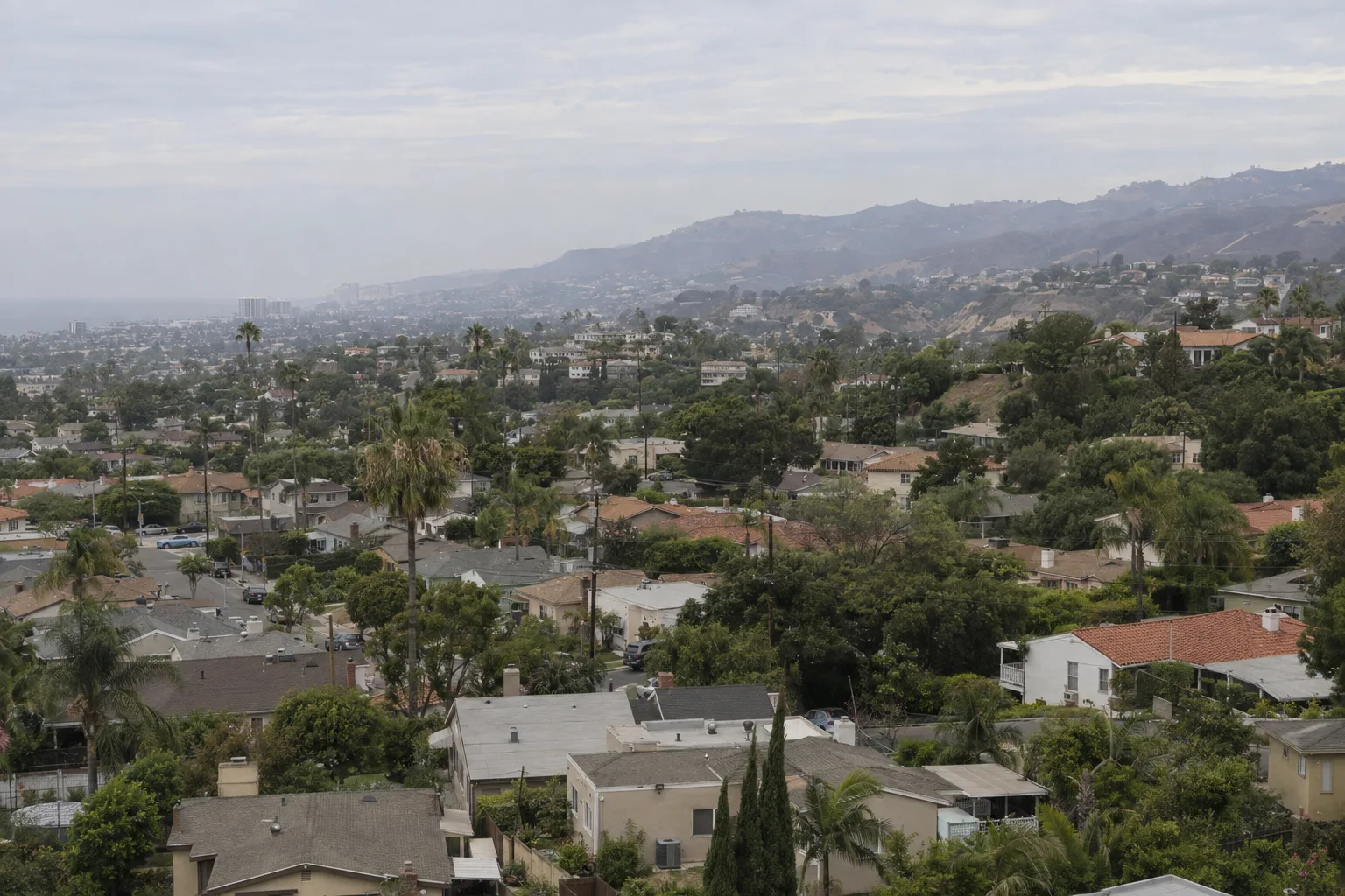 Aerial view of a Los Angeles residential landscape at golden hour showing stucco homes with tile roofs, side-yard AC condensers, palm trees, and hillside terrain illustrating the region's varied climate zones