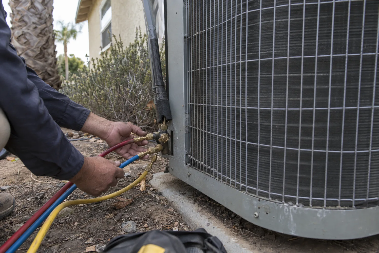 Refrigerant manifold gauges connected to the service ports of a residential central air conditioner condenser unit alongside a stucco house with dry landscaping and a palm tree in the background