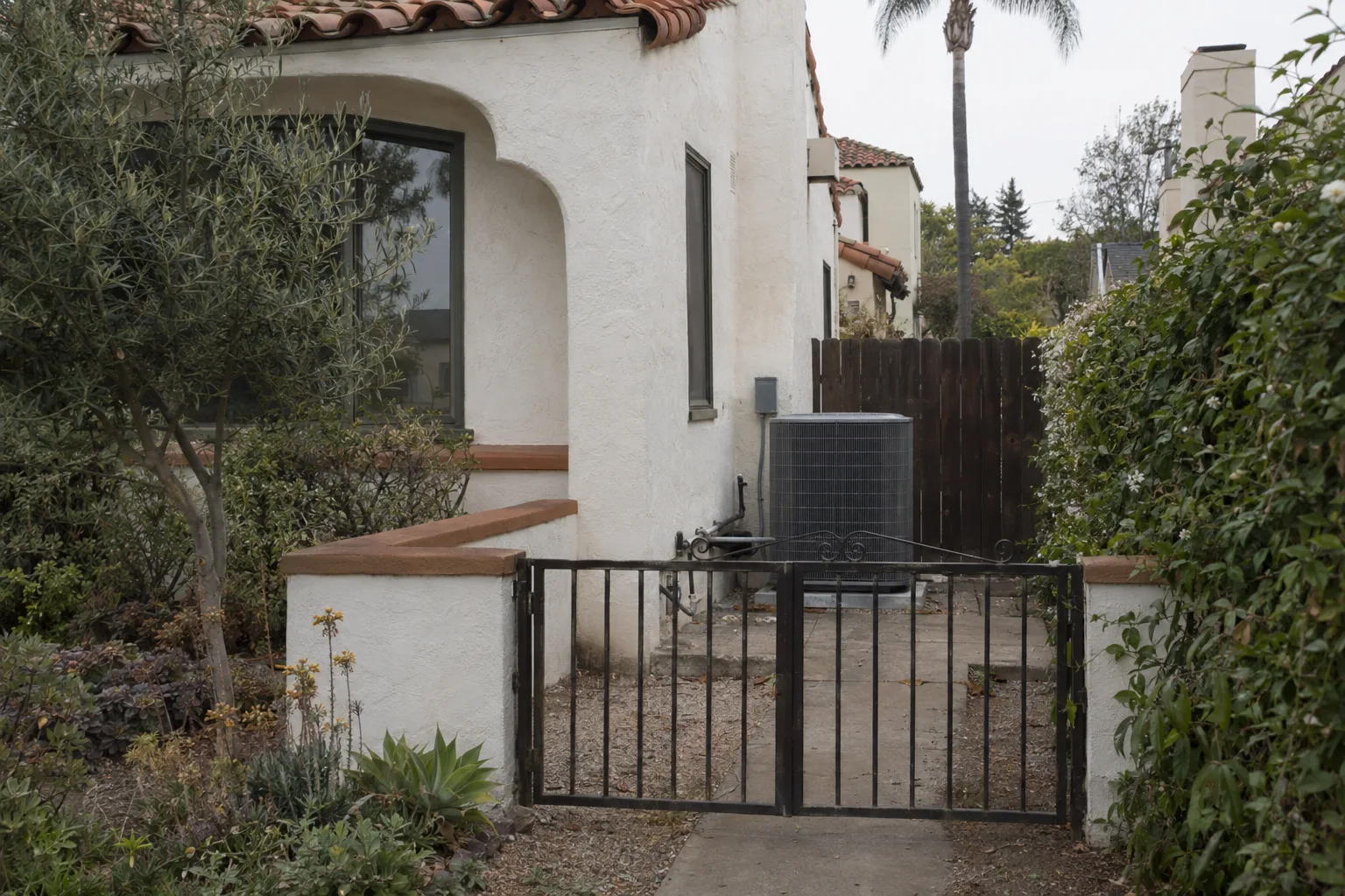 Stucco Spanish-style Los Angeles home exterior at golden hour with a central air conditioning condenser unit installed in the side yard, surrounded by drought-tolerant plants and a tile roof visible above