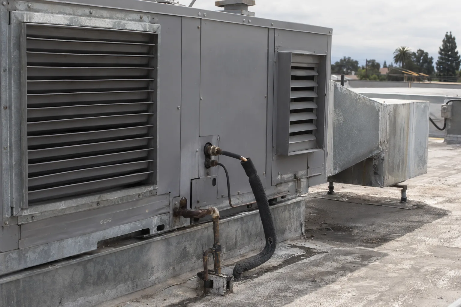 Close-up of the louvered ductwork collar and refrigerant line connections on the side of a rooftop-mounted residential package unit HVAC cabinet, with sunlight highlighting the metal panel seams and hardware.