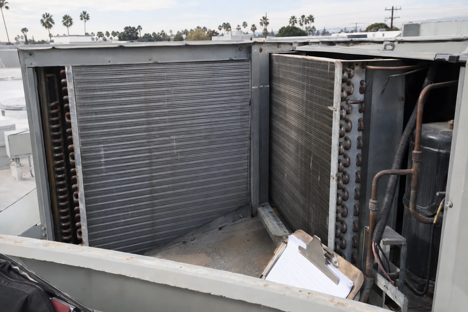 The open interior of a rooftop HVAC package unit showing evaporator and condenser coil surfaces with aluminium fins, with a clipboard resting against the cabinet in the foreground