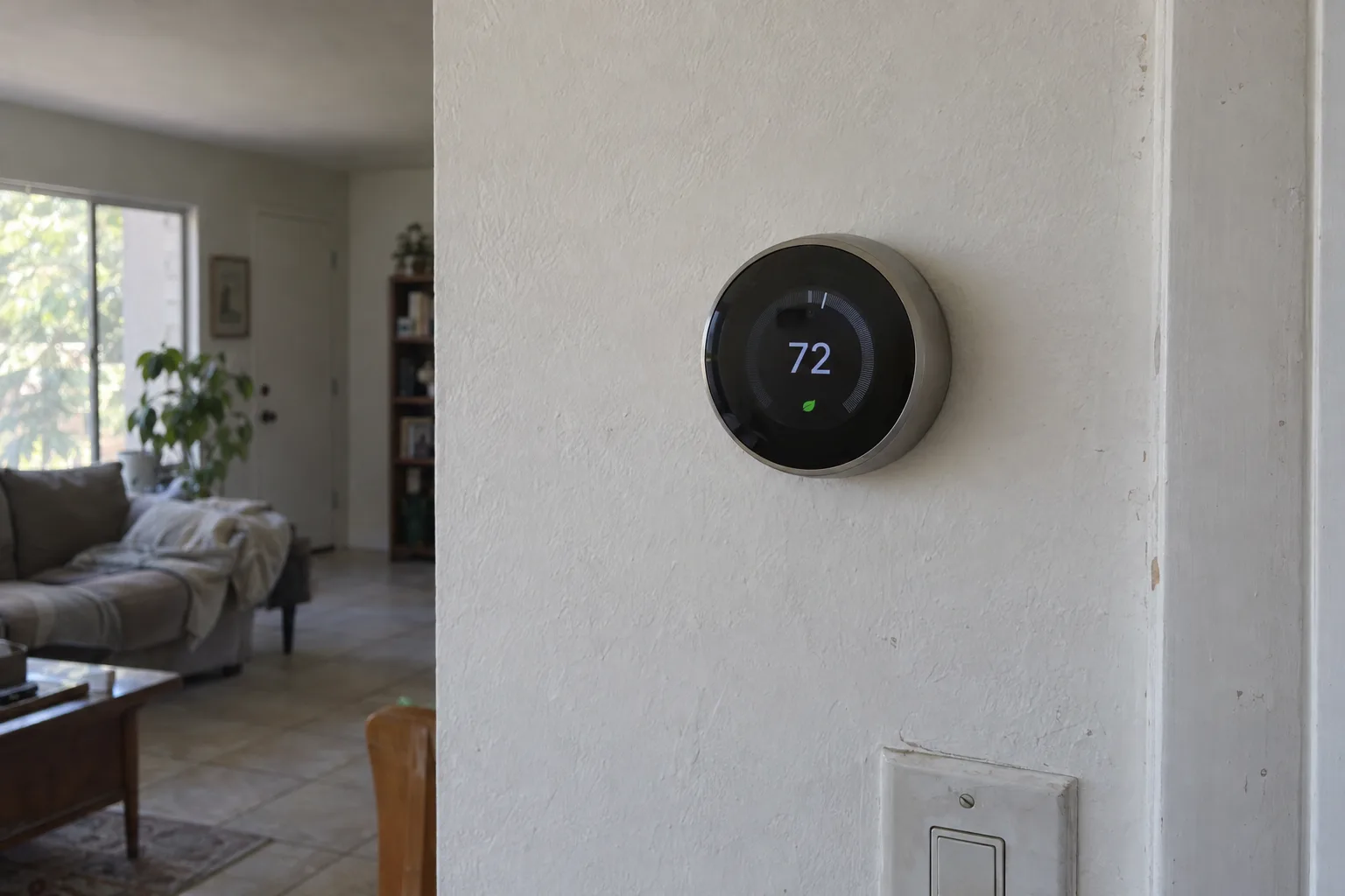 A smart thermostat mounted on a white stucco wall inside a Los Angeles home, its screen glowing with temperature information, warm afternoon light coming from a nearby window