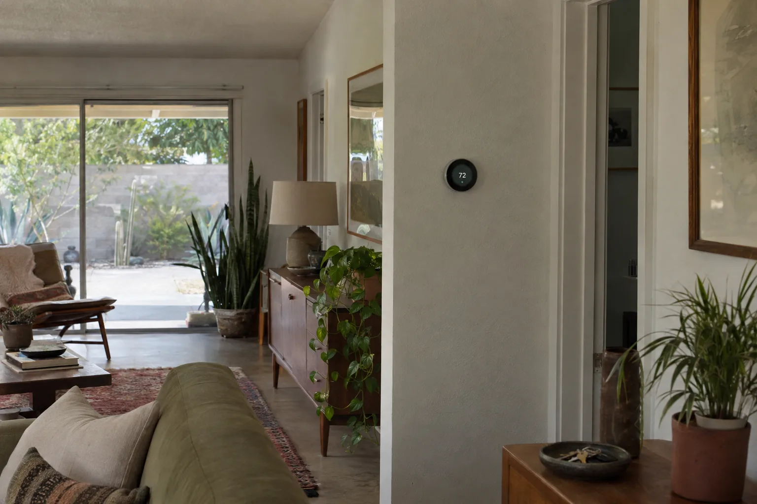 A slim smart thermostat mounted cleanly on a white stucco wall inside a mid-century ranch-style Los Angeles living room, with warm afternoon light and desert plants visible in the background.