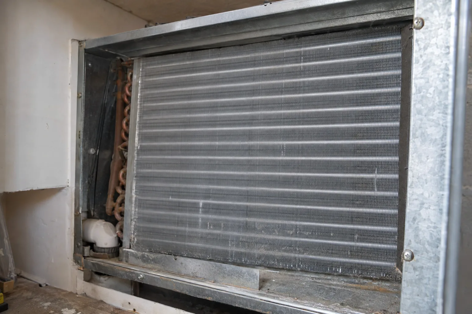 Open access panel on a residential air handler exposing the evaporator coil's dense aluminum fins, lit by a side-angle work light inside a utility closet