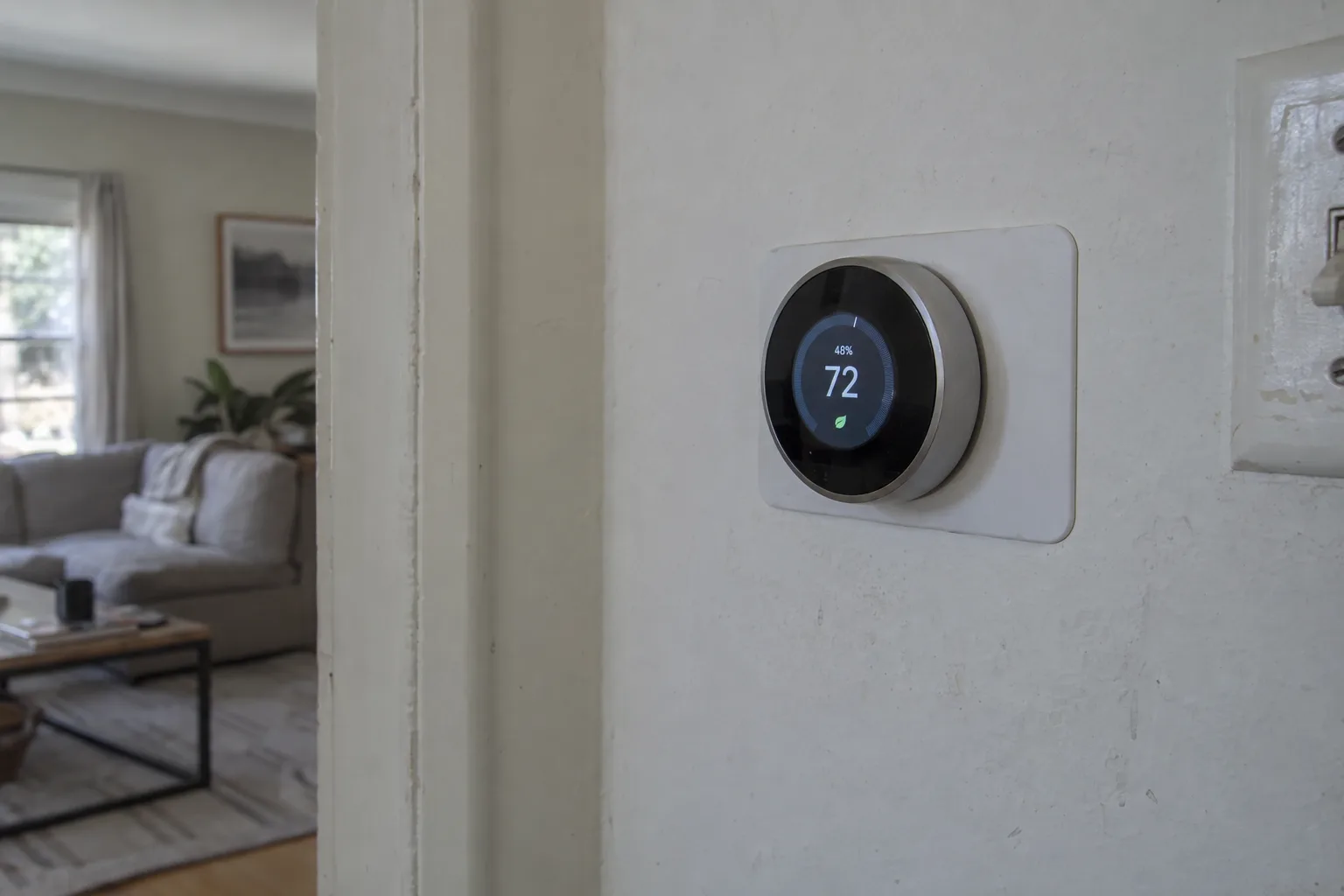 Smart thermostat mounted on an interior wall, its screen glowing with climate readings in soft natural window light