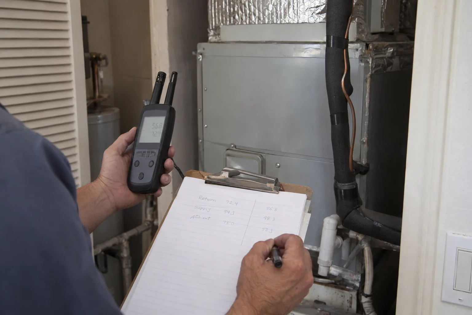 Close-up of a technician's hands holding a digital psychrometer and a clipboard with measurements next to a residential air handler unit inside a utility closet