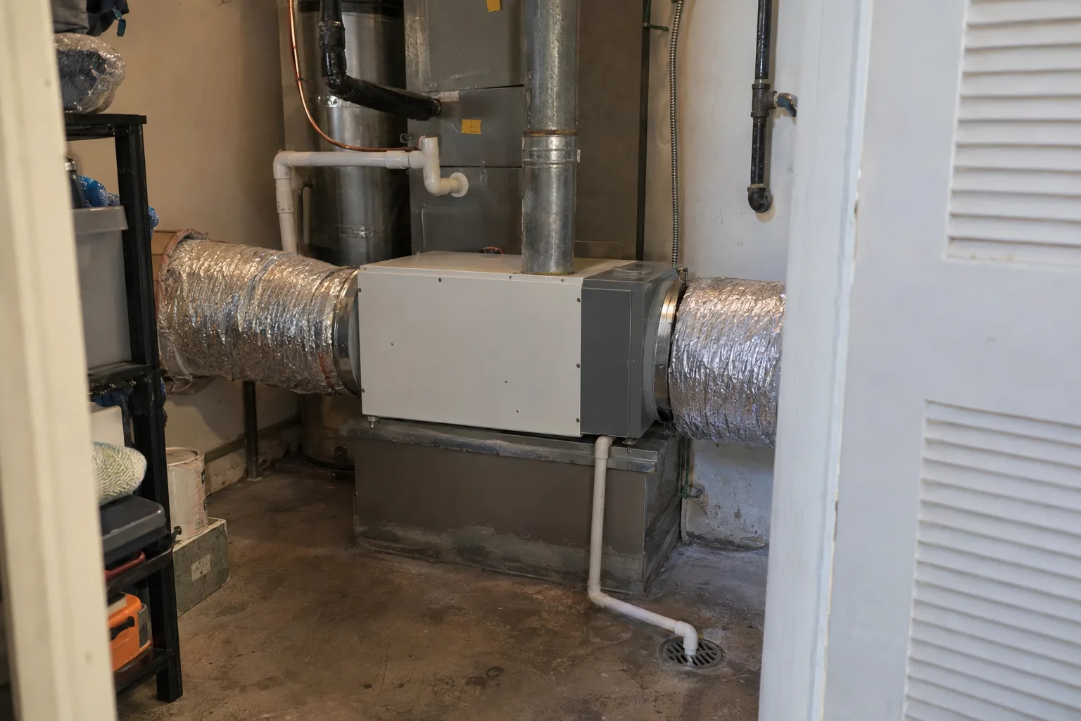 A whole-house dehumidifier mounted inline between ductwork sections inside a residential utility closet, with a condensate drain line routed to a nearby floor drain