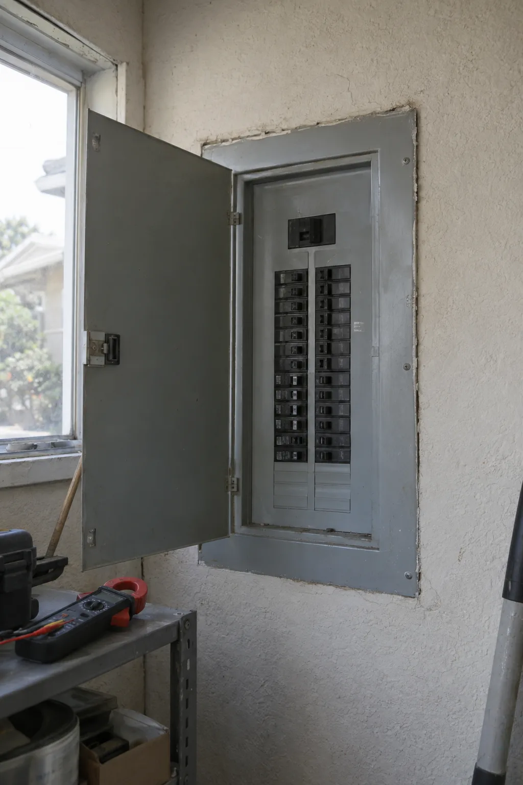 An open residential electrical panel on a stucco interior wall with a clamp meter resting on a nearby shelf, lit by natural window light