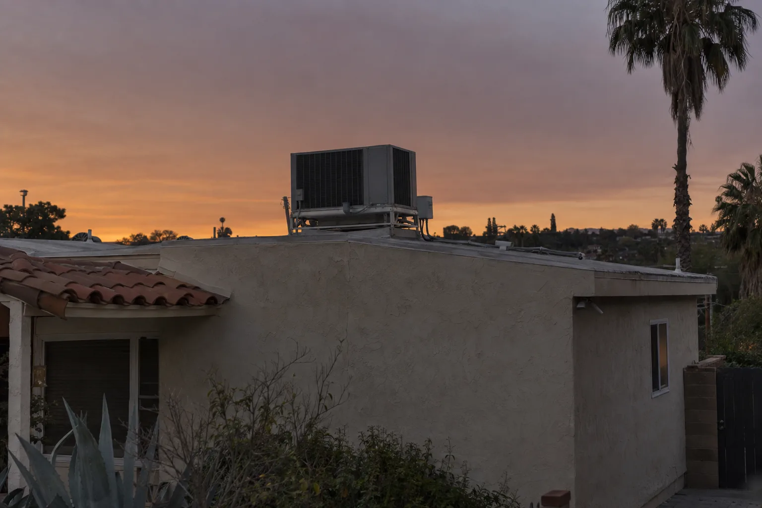 A rooftop air conditioning condenser unit sitting dark and idle on the flat roof of a stucco Los Angeles ranch house at dusk, with a palm tree and fading amber sky in the background.