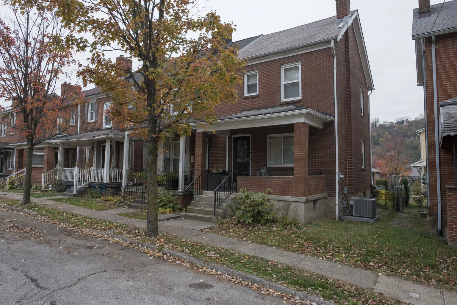 Row of two-story brick rowhouses on a western Pennsylvania residential street with an outdoor AC condenser unit visible beside one home, under overcast skies with mature trees